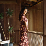 woman with brown hair wearing a brown colored maxi button up dress with light pink lei awapuhi print