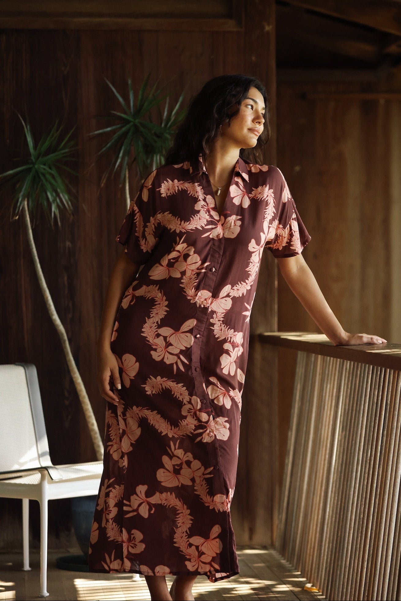 woman with brown hair wearing a brown colored maxi button up dress with light pink lei awapuhi print