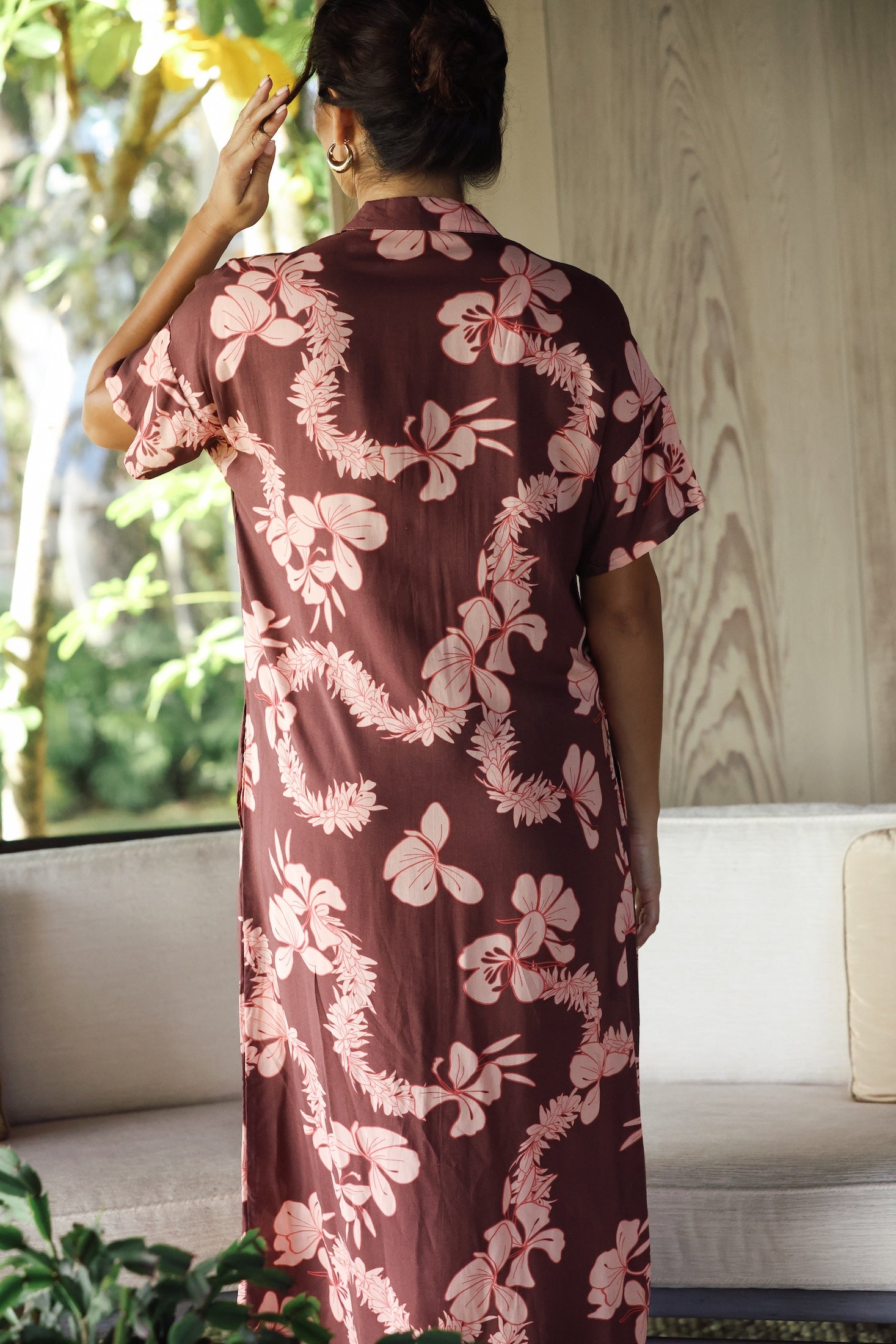 woman with brown hair wearing a brown colored maxi button up dress with light pink lei awapuhi print