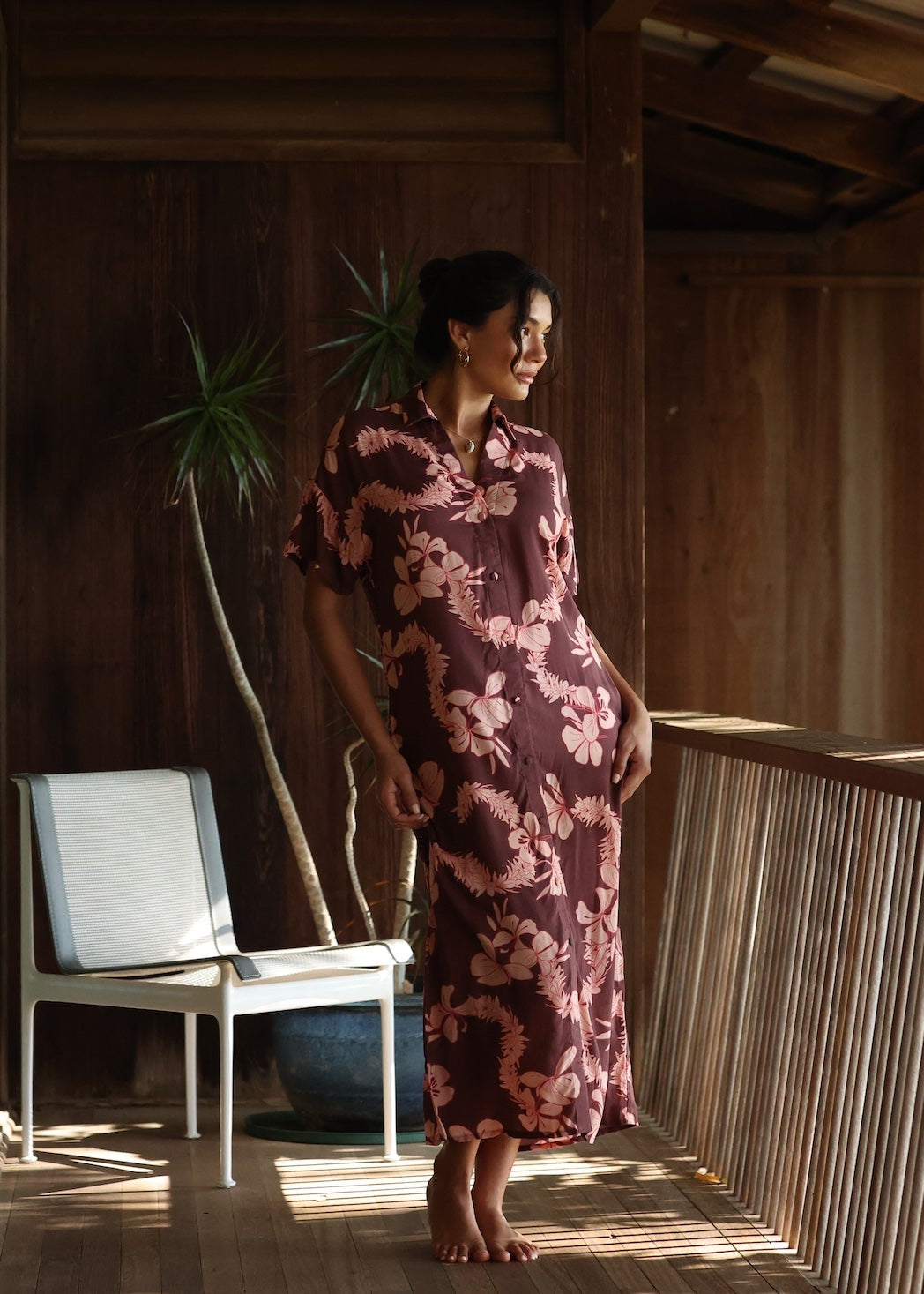 woman with brown hair wearing a brown colored maxi button up dress with light pink lei awapuhi print