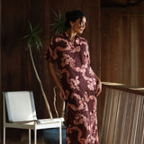 woman with brown hair wearing a brown colored maxi button up dress with light pink lei awapuhi print