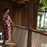 woman with brown hair wearing a brown colored maxi button up dress with light pink lei awapuhi print