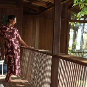 woman with brown hair wearing a brown colored maxi button up dress with light pink lei awapuhi print