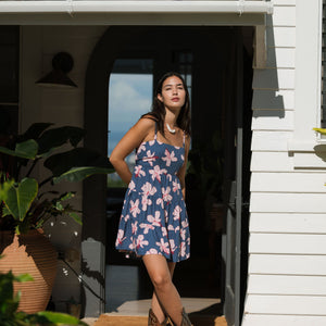 woman with black hair wearing a mini tiered dress with tie back in a navy color with pink hibiscus blossoms