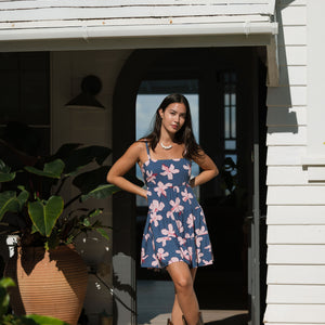 woman with black hair wearing a mini tiered dress with tie back in a navy color with pink hibiscus blossoms