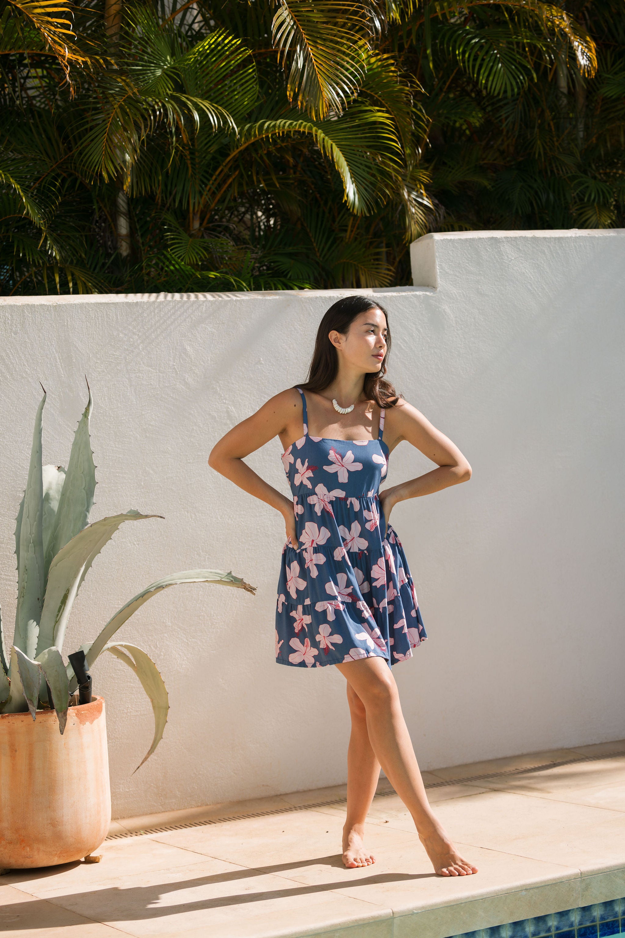 woman with black hair wearing a mini tiered dress with tie back in a navy color with pink hibiscus blossoms