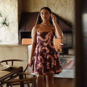 woman with brown hair wearing a brown colored tie back mini dress  with light pink lei awapuhi print