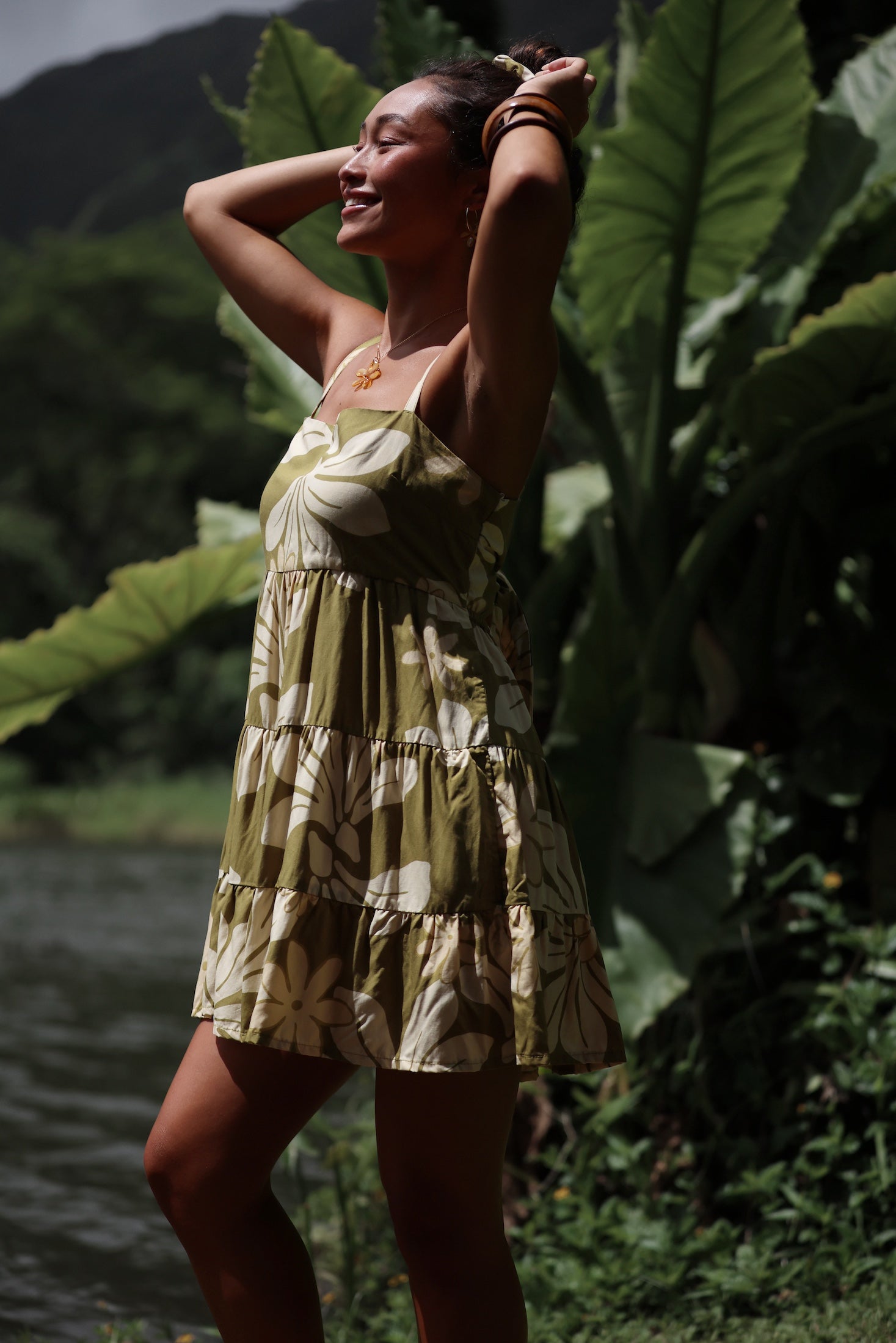 woman with brown hair wearing a mini dress with a tie back in a green color with naupaka pattern throughout.