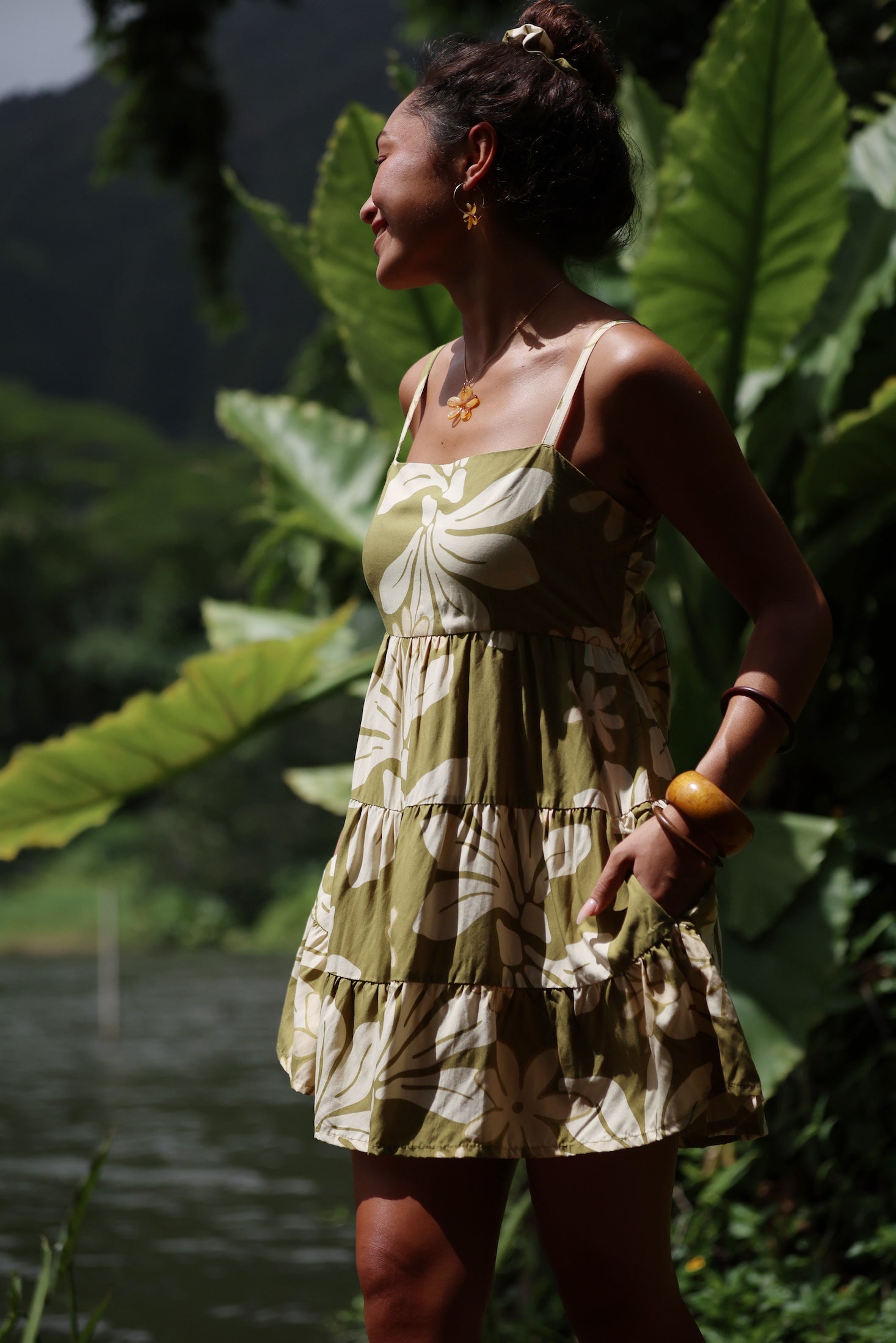 woman with brown hair wearing a mini dress with a tie back in a green color with naupaka pattern throughout.