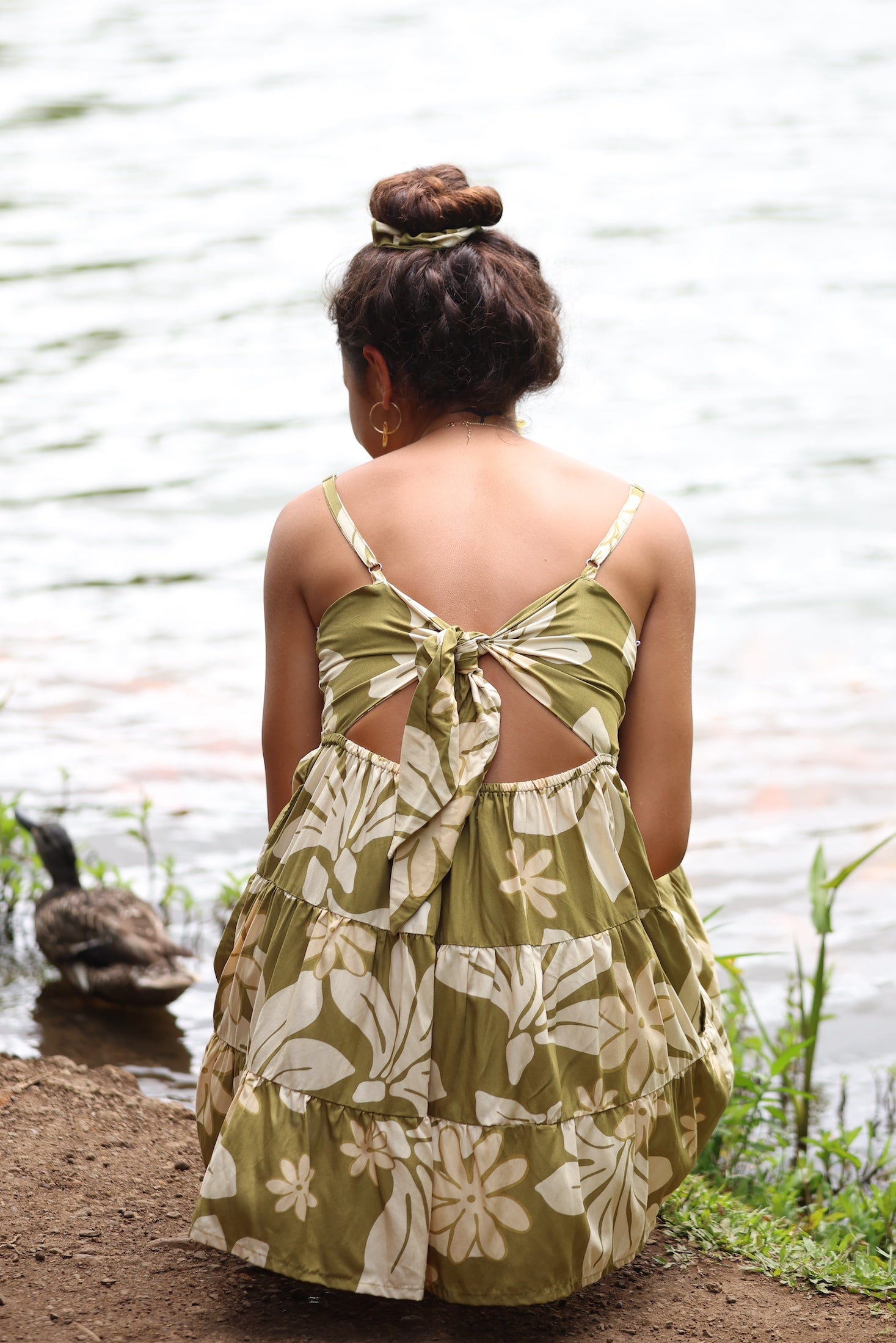 woman with brown hair wearing a mini dress with a tie back in a green color with naupaka pattern throughout.