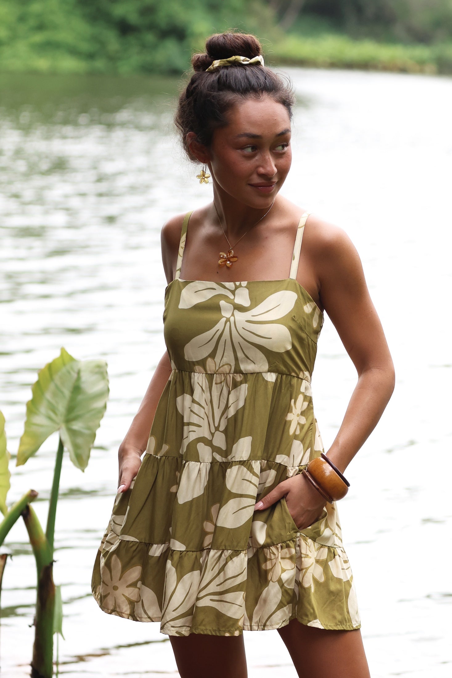 woman with brown hair wearing a mini dress with a tie back in a green color with naupaka pattern throughout.