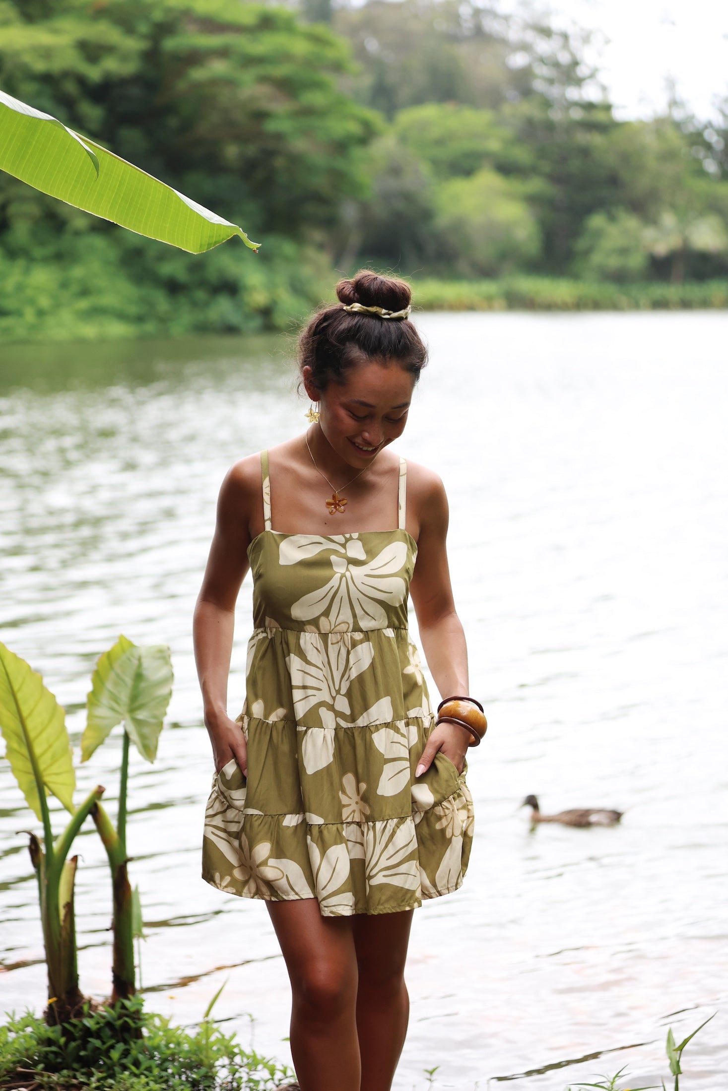 woman with brown hair wearing a mini dress with a tie back in a green color with naupaka pattern throughout.