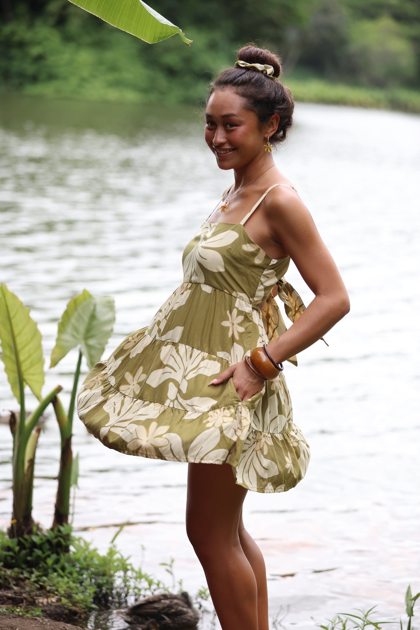 woman with brown hair wearing a mini dress with a tie back in a green color with naupaka pattern throughout.