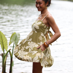 woman with brown hair wearing a mini dress with a tie back in a green color with naupaka pattern throughout.