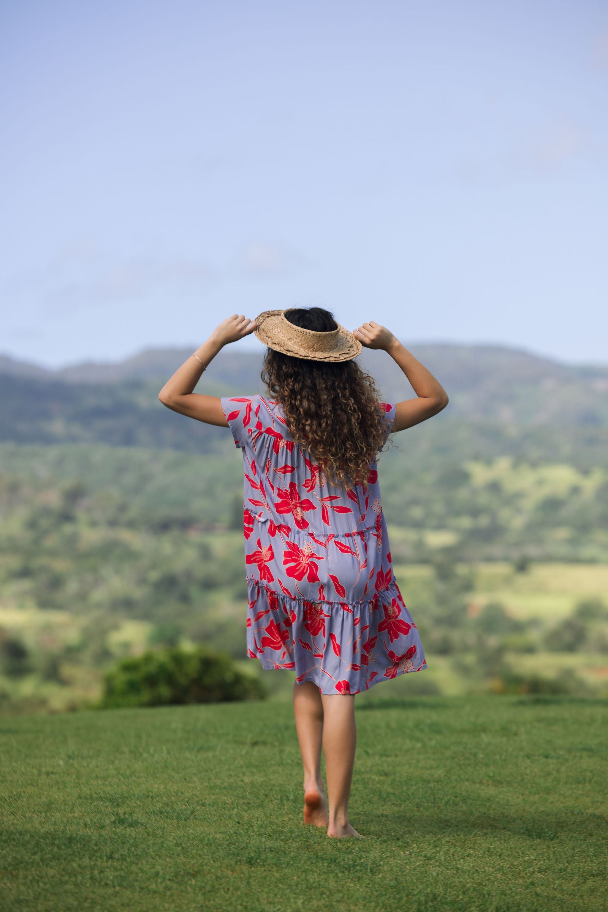 woman with brown hair wearing a flowy dress with periwinkle background and bright red hibiscus print.