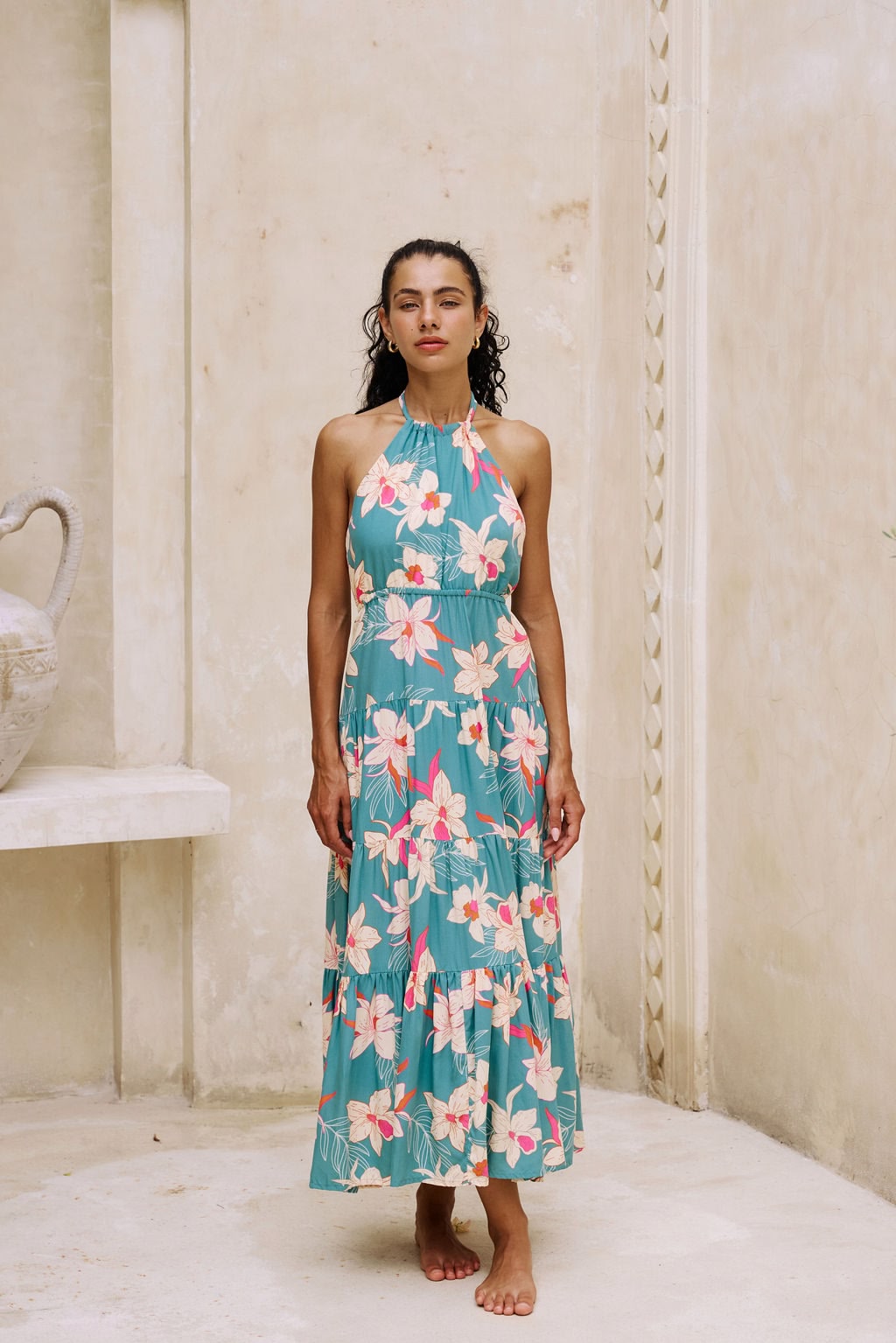 woman with brown hair wearing tiered halter maxi dress in teal with large cream colored orchids.
