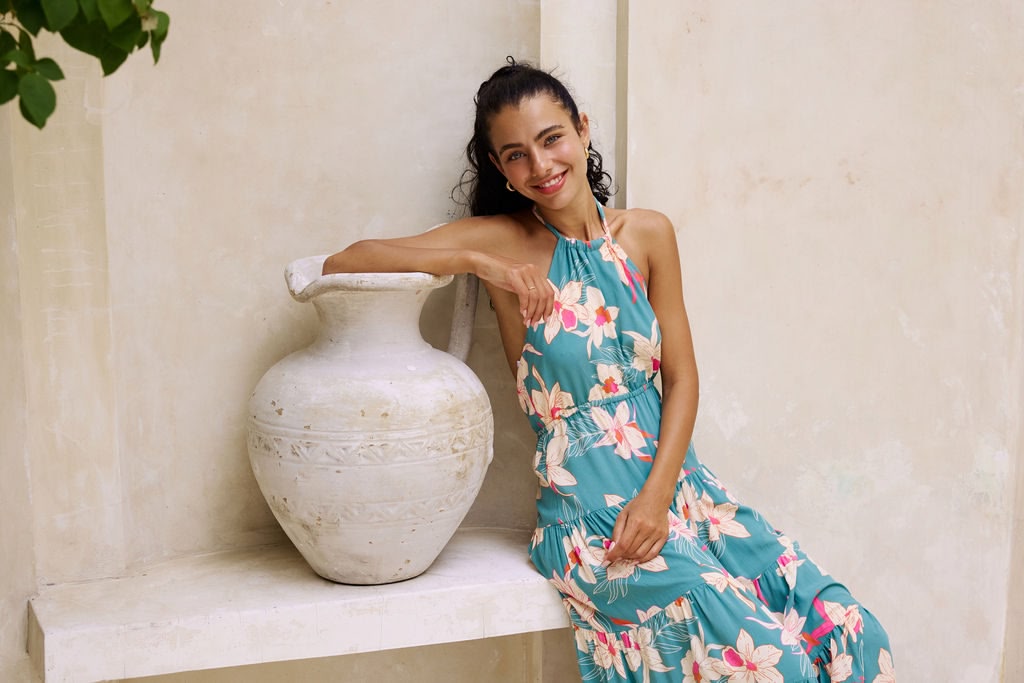 woman with brown hair wearing tiered halter maxi dress in teal with large cream colored orchids.
