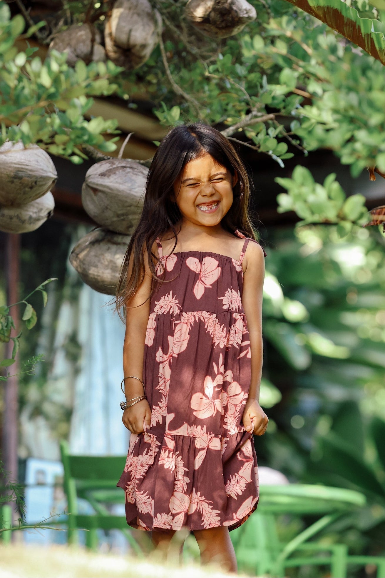 little girl wearing a long brown dress with tie sleeve with light pink lei awapuhi print