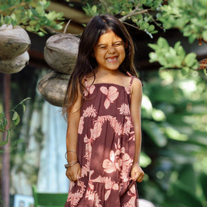 little girl wearing a long brown dress with tie sleeve with light pink lei awapuhi print