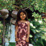 little girl wearing a long brown dress with tie sleeve with light pink lei awapuhi print
