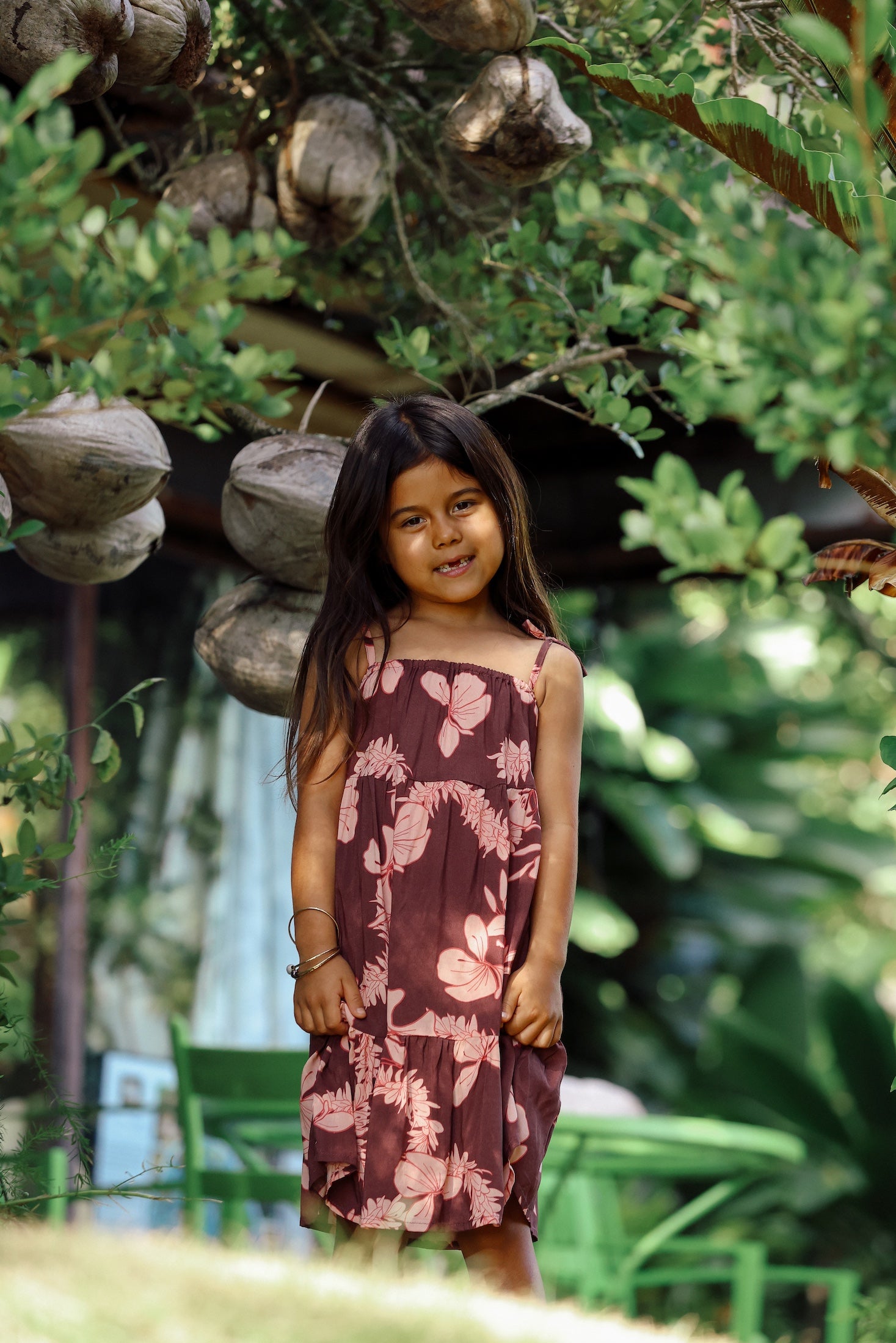 little girl wearing a long brown dress with tie sleeve with light pink lei awapuhi print