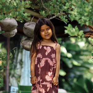 little girl wearing a long brown dress with tie sleeve with light pink lei awapuhi print