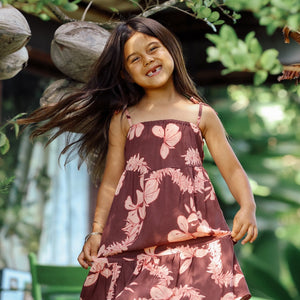 little girl wearing a long brown dress with tie sleeve with light pink lei awapuhi print