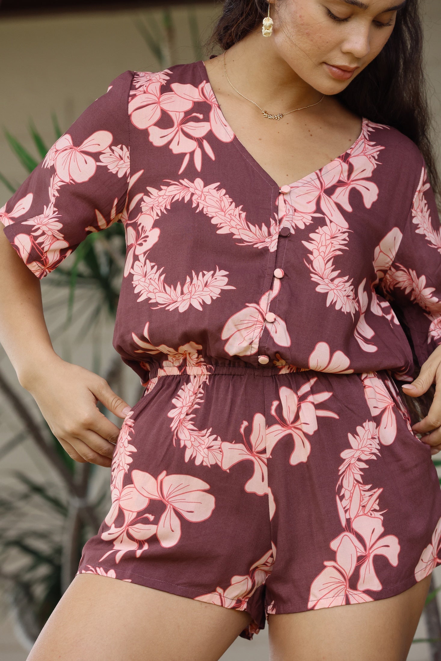 woman with brown hair wearing brown button up playsuit with light pink lei awapuhi print