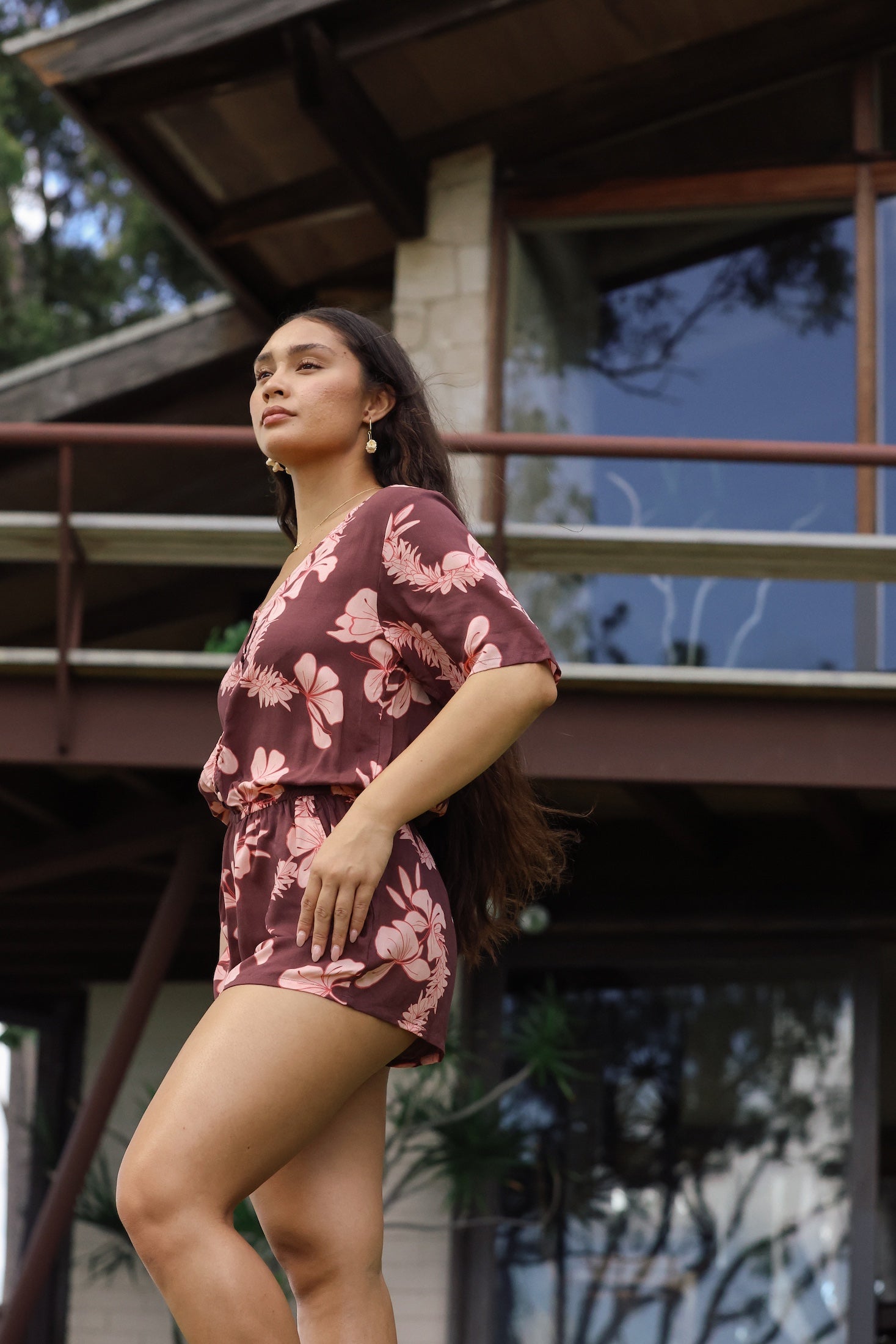 woman with brown hair wearing brown button up playsuit with light pink lei awapuhi print