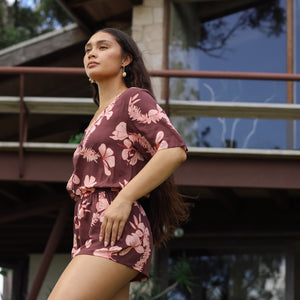 woman with brown hair wearing brown button up playsuit with light pink lei awapuhi print