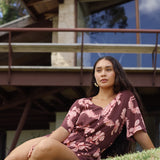 woman with brown hair wearing brown button up playsuit with light pink lei awapuhi print