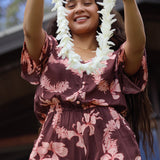 woman with brown hair wearing brown button up playsuit with light pink lei awapuhi print