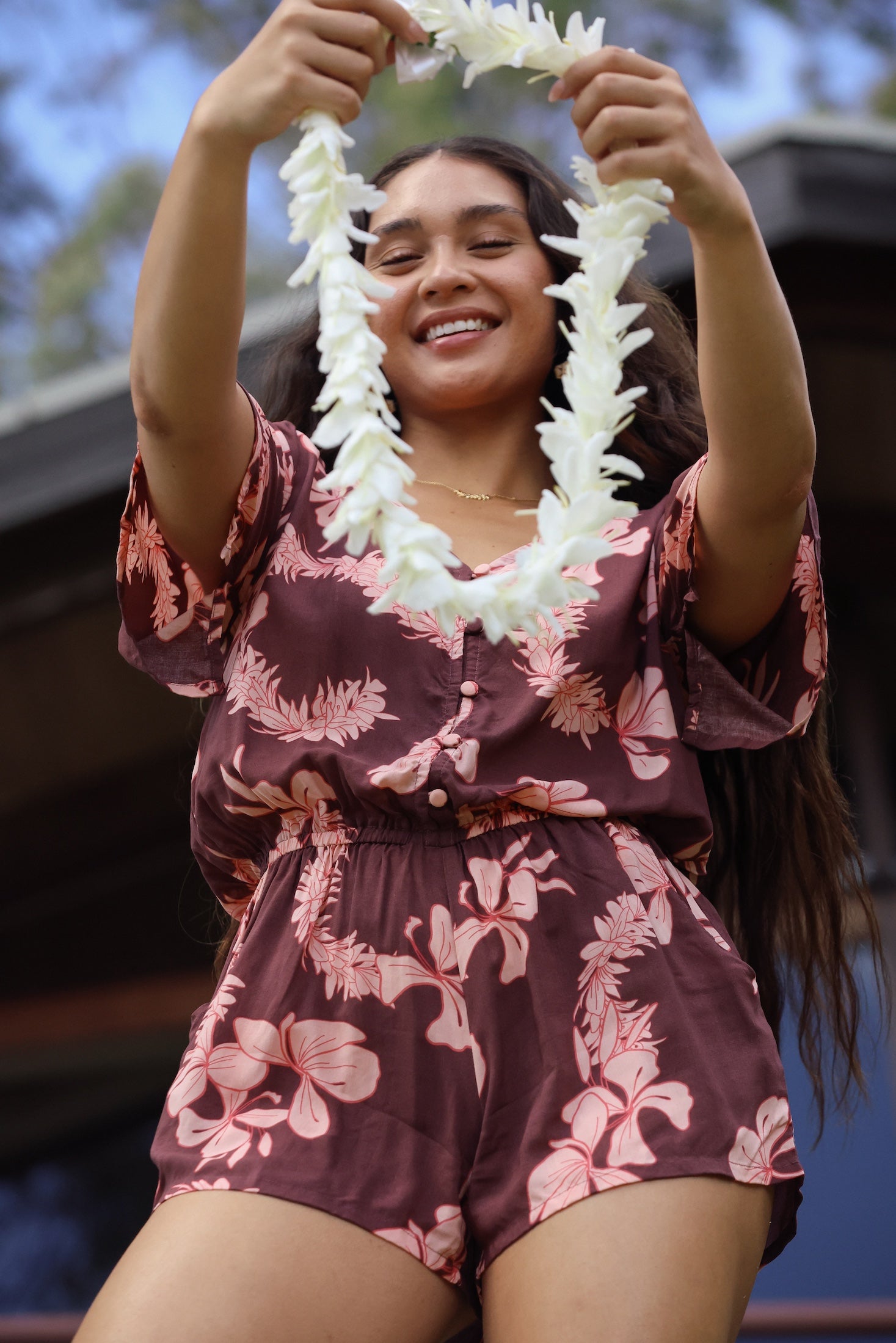 woman with brown hair wearing brown button up playsuit with light pink lei awapuhi print
