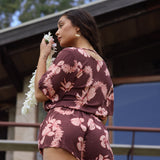 woman with brown hair wearing brown button up playsuit with light pink lei awapuhi print