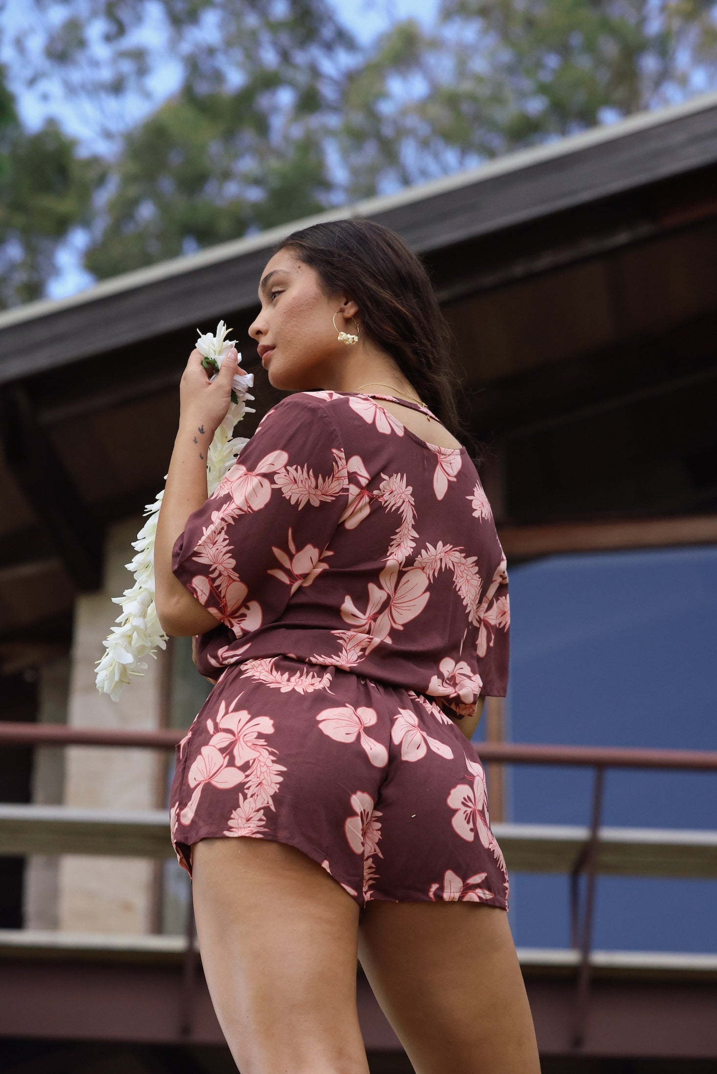 woman with brown hair wearing brown button up playsuit with light pink lei awapuhi print