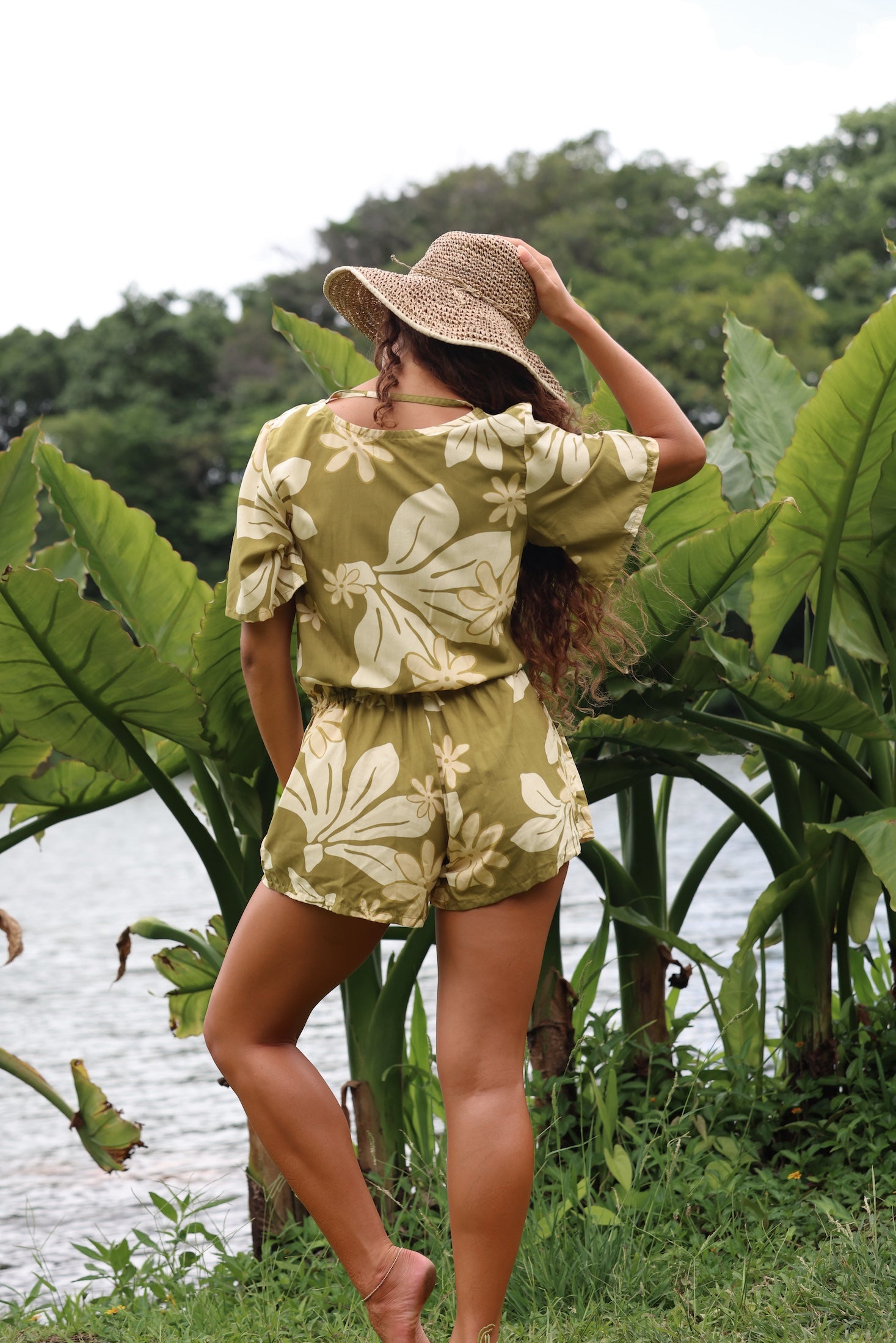 woman with brown hair wearing a short sleeve playsuit in a green color with naupaka pattern throughout.