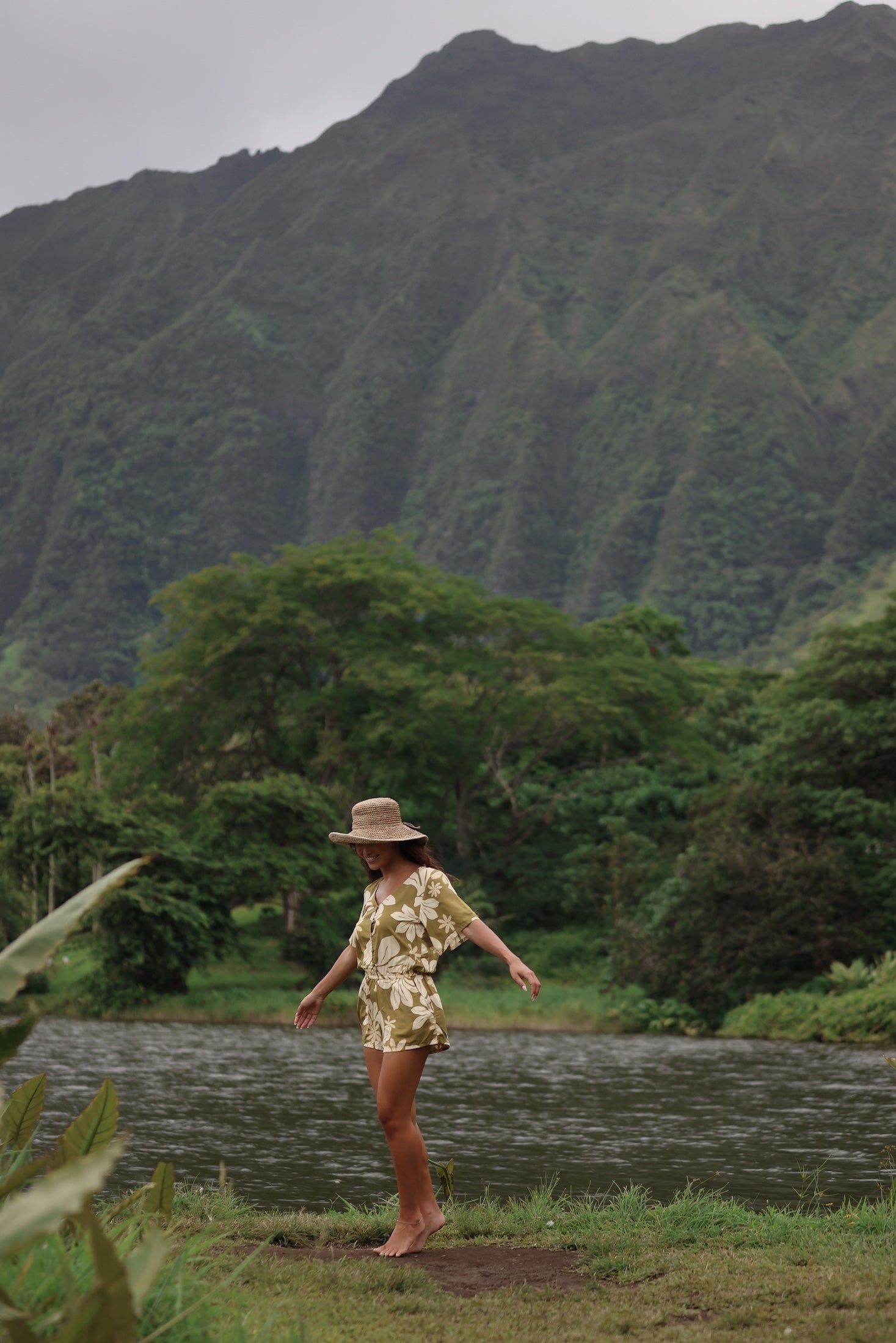 woman with brown hair wearing a short sleeve playsuit in a green color with naupaka pattern throughout.