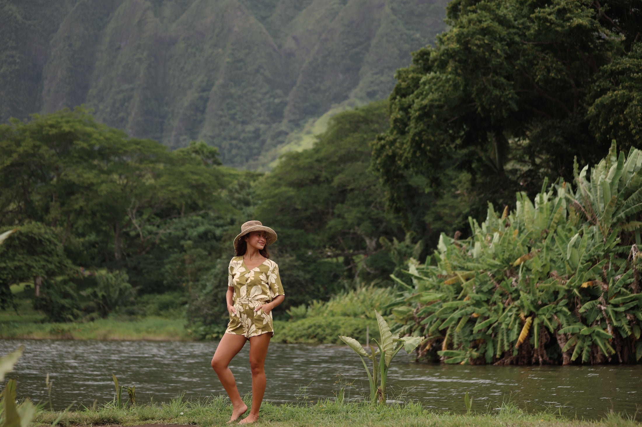 woman with brown hair wearing a short sleeve playsuit in a green color with naupaka pattern throughout.