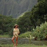 woman with brown hair wearing a short sleeve playsuit in a green color with naupaka pattern throughout.