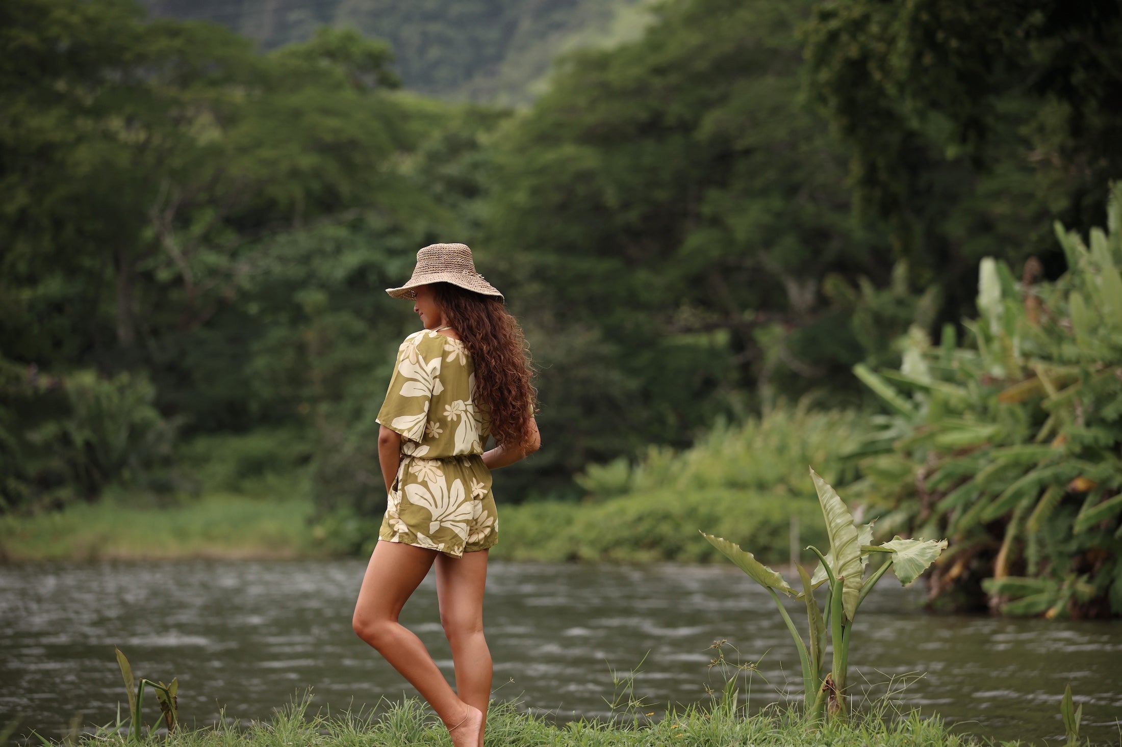 woman with brown hair wearing a short sleeve playsuit in a green color with naupaka pattern throughout.