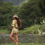 woman with brown hair wearing a short sleeve playsuit in a green color with naupaka pattern throughout.