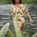 woman with brown hair wearing a short sleeve playsuit in a green color with naupaka pattern throughout.
