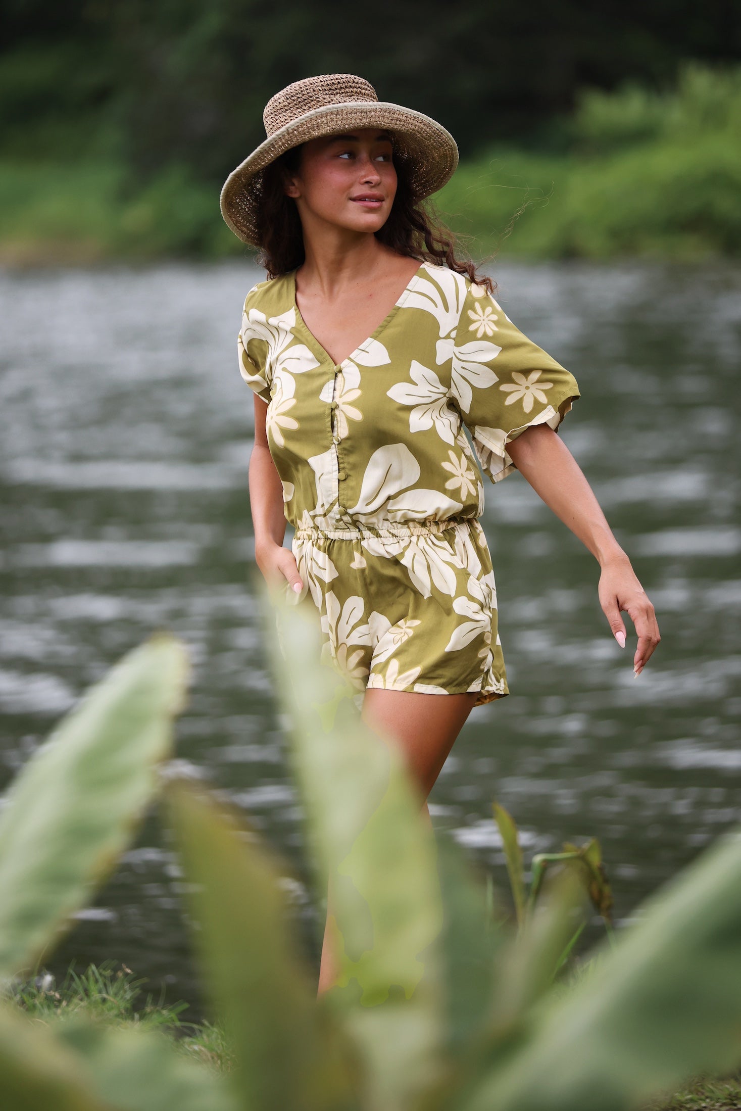 woman with brown hair wearing a short sleeve playsuit in a green color with naupaka pattern throughout.