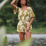 woman with brown hair wearing a short sleeve playsuit in a green color with naupaka pattern throughout.
