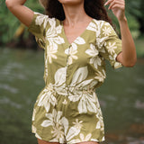 woman with brown hair wearing a short sleeve playsuit in a green color with naupaka pattern throughout.