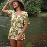 woman with brown hair wearing a short sleeve playsuit in a green color with naupaka pattern throughout.