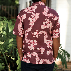 man wearing brown aloha shirt with pink lei awapuhi print