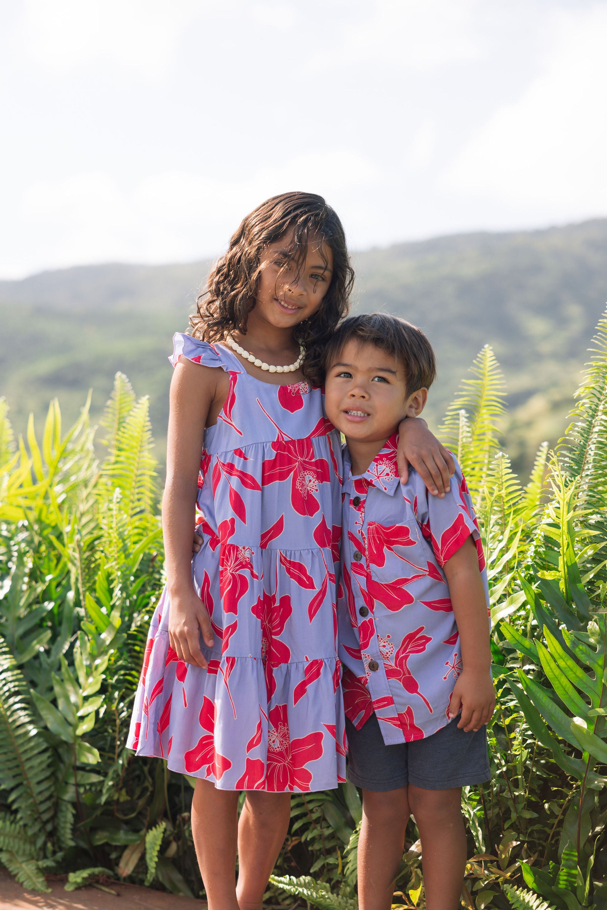 little girl with brown hair wearing a mid length dress with periwinkle background and bright red hibiscus print.