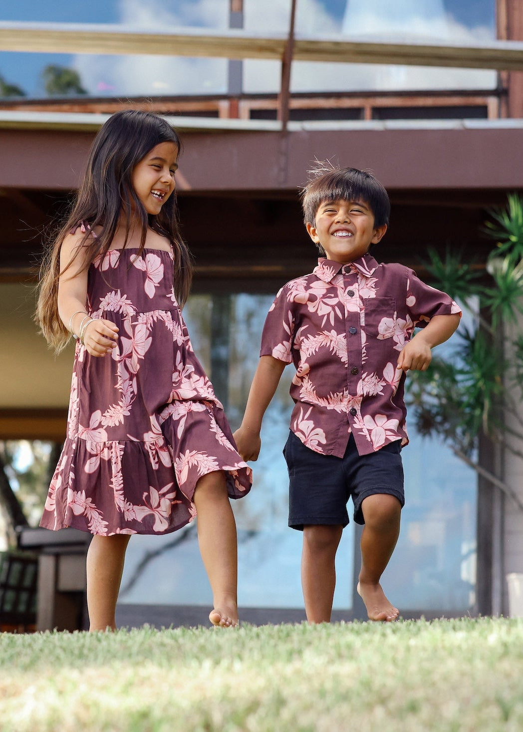 little girl wearing a long brown dress with tie sleeve with light pink lei awapuhi print and little boy wearing brown button-up with light pink lei awapuhi print