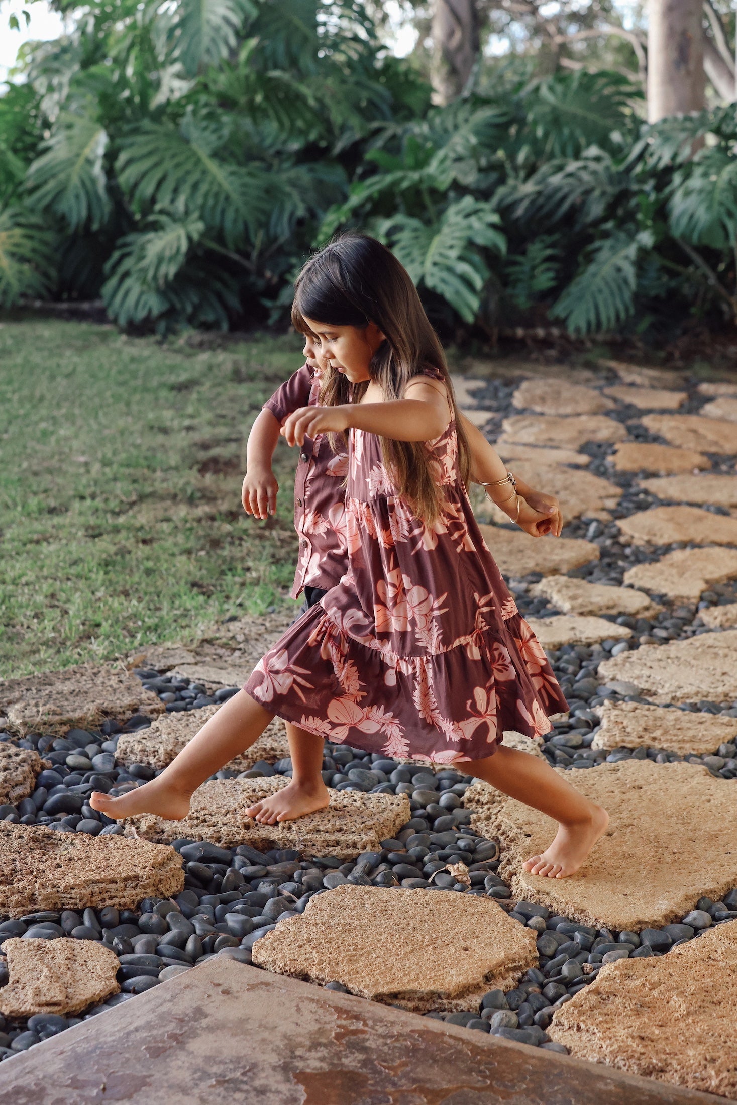 little girl wearing a long brown dress with tie sleeve with light pink lei awapuhi print